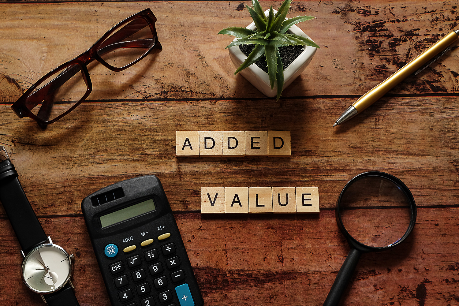 Top view wood text Added value with brown glasses,calculator,gold pen,black watch,magnifying glass and white pot plant.